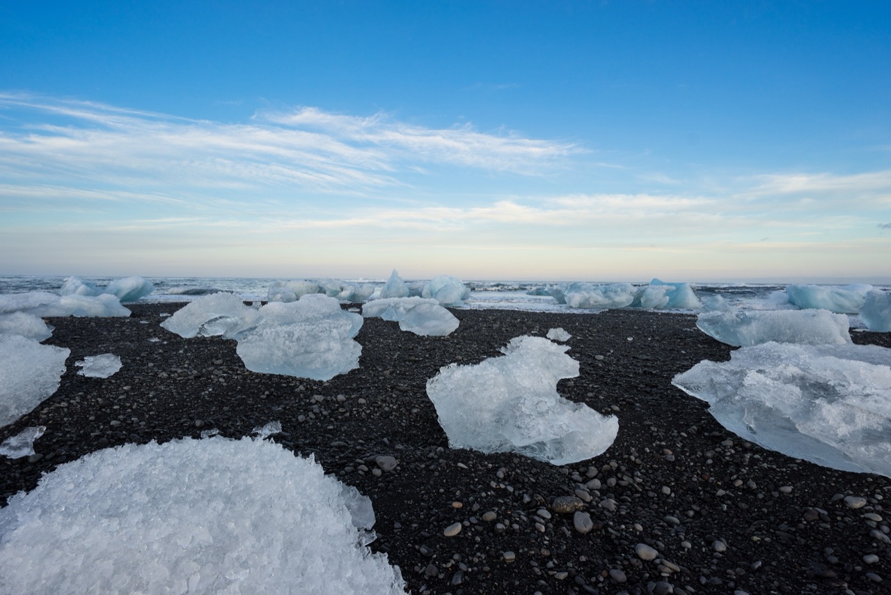 Blocks of clear ice scattered across wet black volcanic sand under a bright blue sky.
