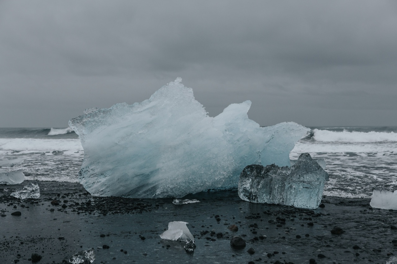 A large weathered block of ice stranded on black sand at the tideline, with surf breaking behind it.