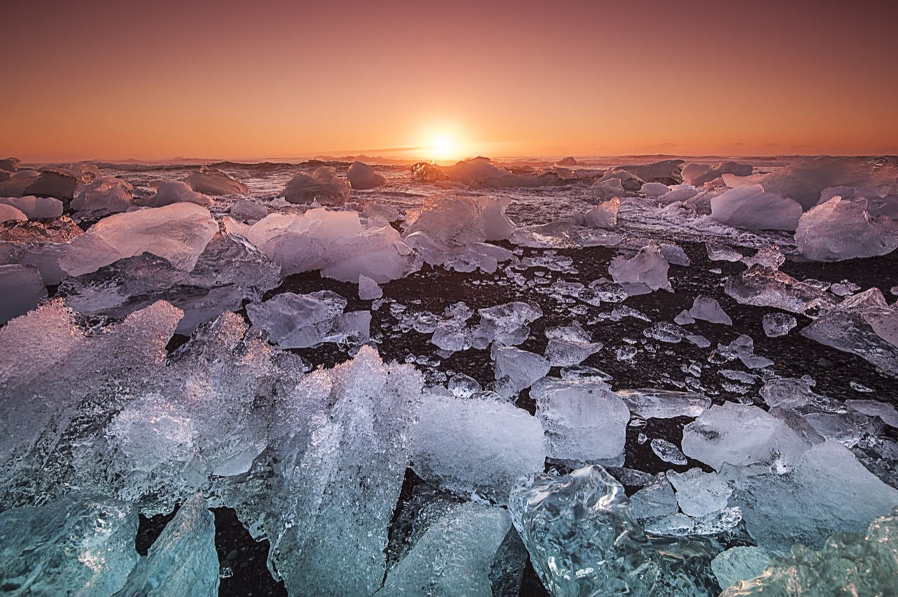 Low sun burning orange on the horizon behind a shoreline packed with fragments of ice.