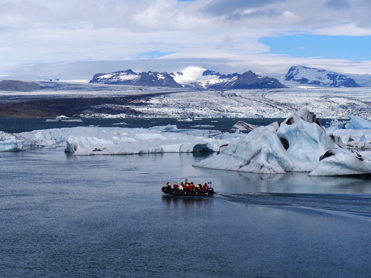 A small inflatable boat carrying passengers in orange flotation suits, dwarfed by icebergs on the lagoon with snow-covered mountains behind.